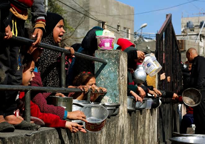 A picture of hungry women and kids asking for food with their plates through a gate 