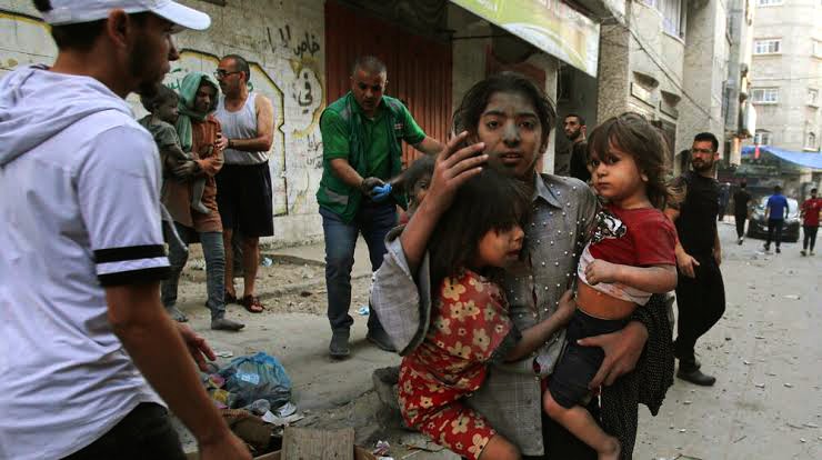 The Dire Humanitarian Situation in Gaza: A picture of a woman looking unkempt while holding two children on both hands,  standing in a street with people 