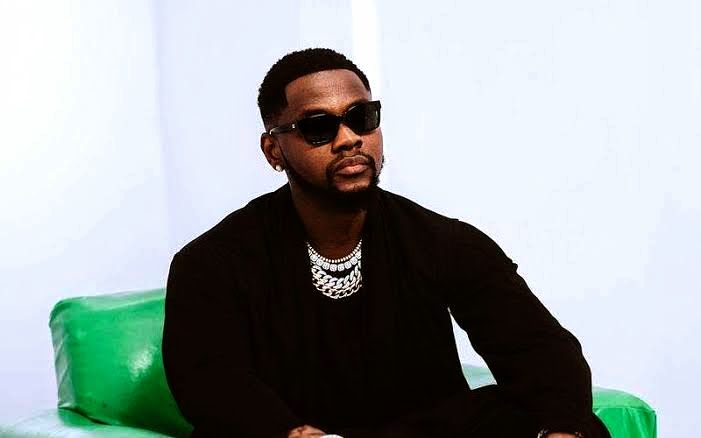 A picture of Kizz Daniel dressed in an all-black outfit, putting on a black eye shield and ice-chains on his neck while sitting on a green chair