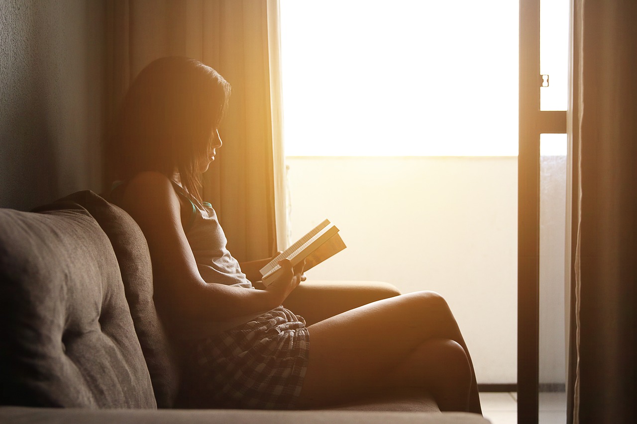 A picture of a young lady sitting on a couch while studying. The area is well-lit with natural light streaming in from a nearby window